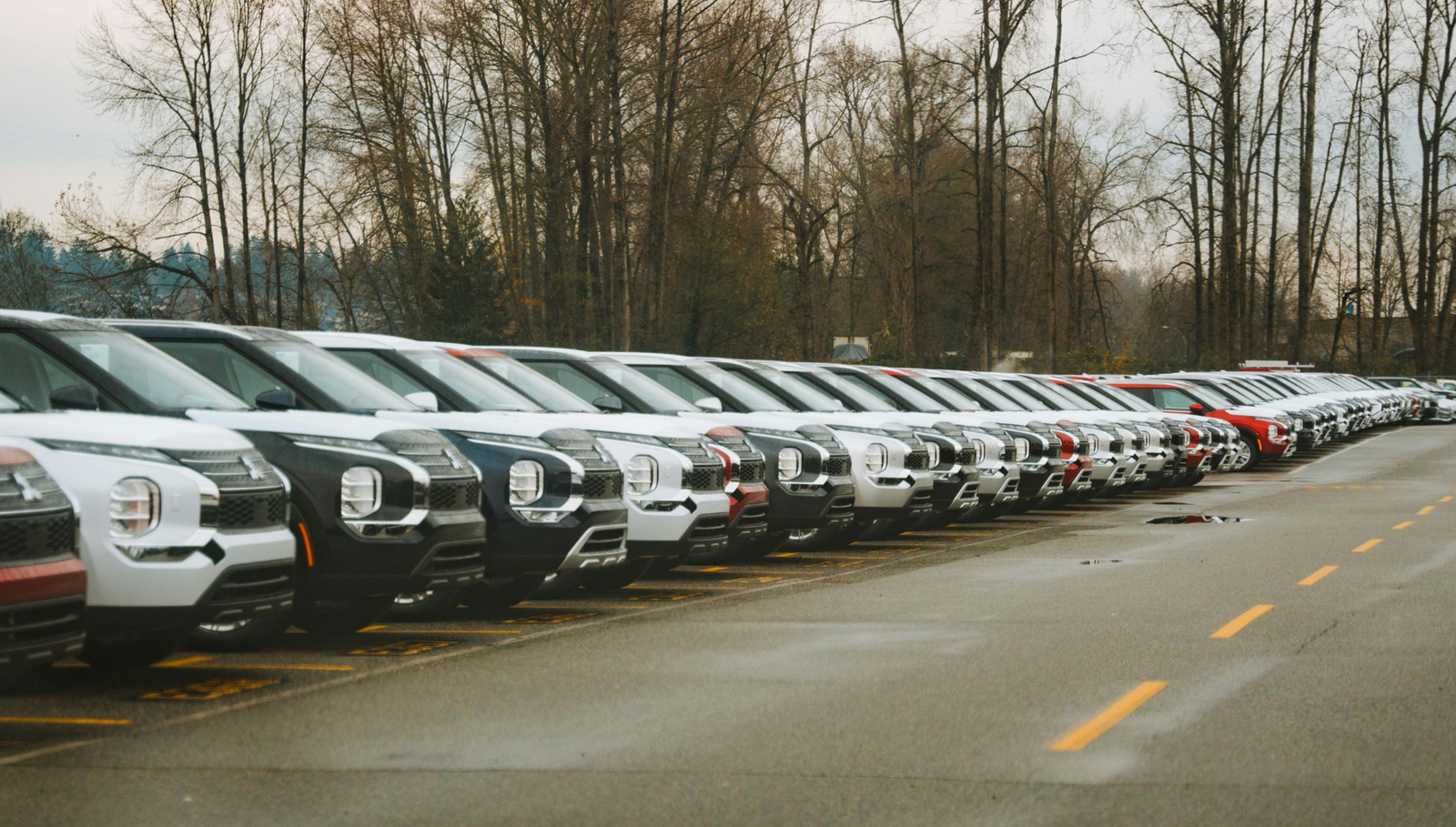 Row of new compact SUVs parked in a dealership lot on a cloudy day with bare trees in the background.